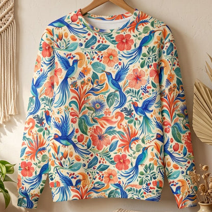 A long-sleeve crewneck sweatshirt featuring a vibrant, all-over Mexican-style print with blue birds, red flowers, and green foliage on an off-white background, hanging on a wooden hanger against a textured beige wall.