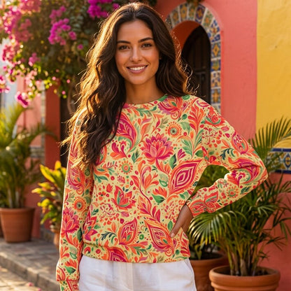 A woman stands on a cobblestone street wearing a long-sleeve crewneck top with a vibrant pink, red, and green floral paisley pattern on a yellow-orange background, paired with white shorts.